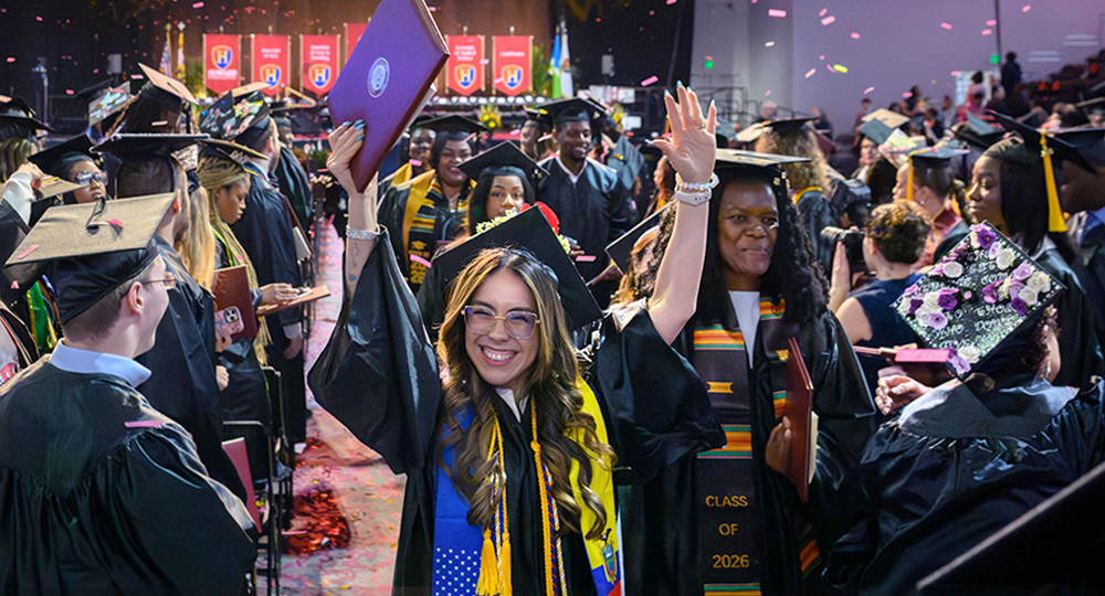 Howard Community College graduates celebrate during a commencement ceremony, smiling and waving in caps and gowns as confetti falls around them.