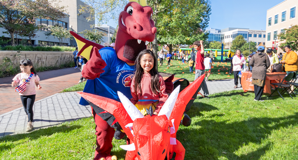 A young girl smiles while posing with Howard Community College’s red dragon mascot during an outdoor campus event. Other families and children enjoy activities on the grassy quad under sunny skies.