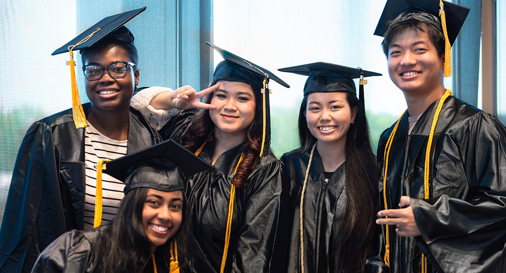 Group of five HCC graduates in black caps and gowns smiling and posing together indoors, with one student making a peace sign.