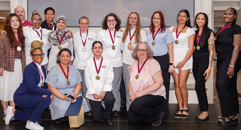 “Group photo of students, faculty, and staff wearing medals at a recognition ceremony, with several culinary students in chef coats posing together indoors.