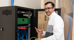 A man wearing glasses and a white jacket with gray sleeves is working on a network cabinet. He is holding a yellow Ethernet cable and smiling at the camera. The cabinet contains multiple blue and red network cables, a switch, and other networking equipment. The background shows exposed wiring and a partially finished wall.