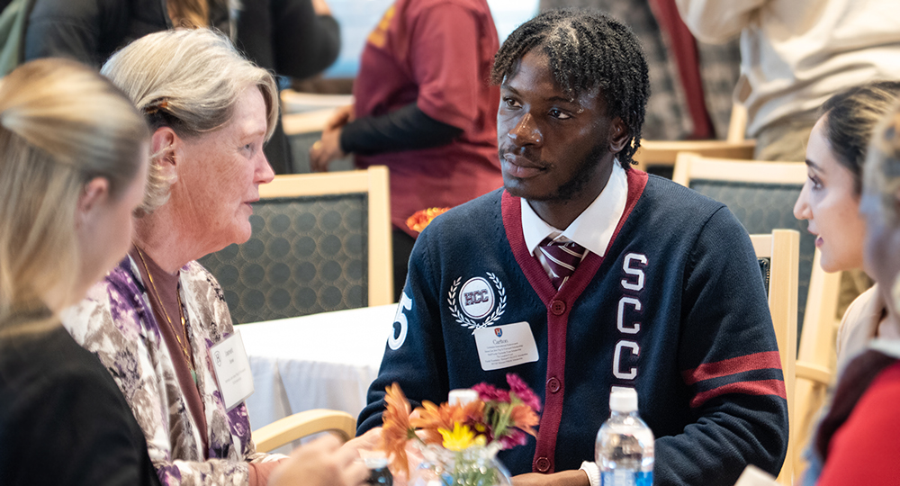 A group of people sit together at a round table during an event. A student wearing a navy cardigan with red accents and college initials listens attentively to an older woman speaking. Others at the table also engage in conversation. A small vase of flowers and a water bottle sit on the table, with other attendees visible in the background.