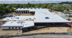 Aerial view of a large building under construction on a college campus, showing a wide roof with several blue-covered sections, exposed framing on one side, and construction equipment and materials surrounding the site.