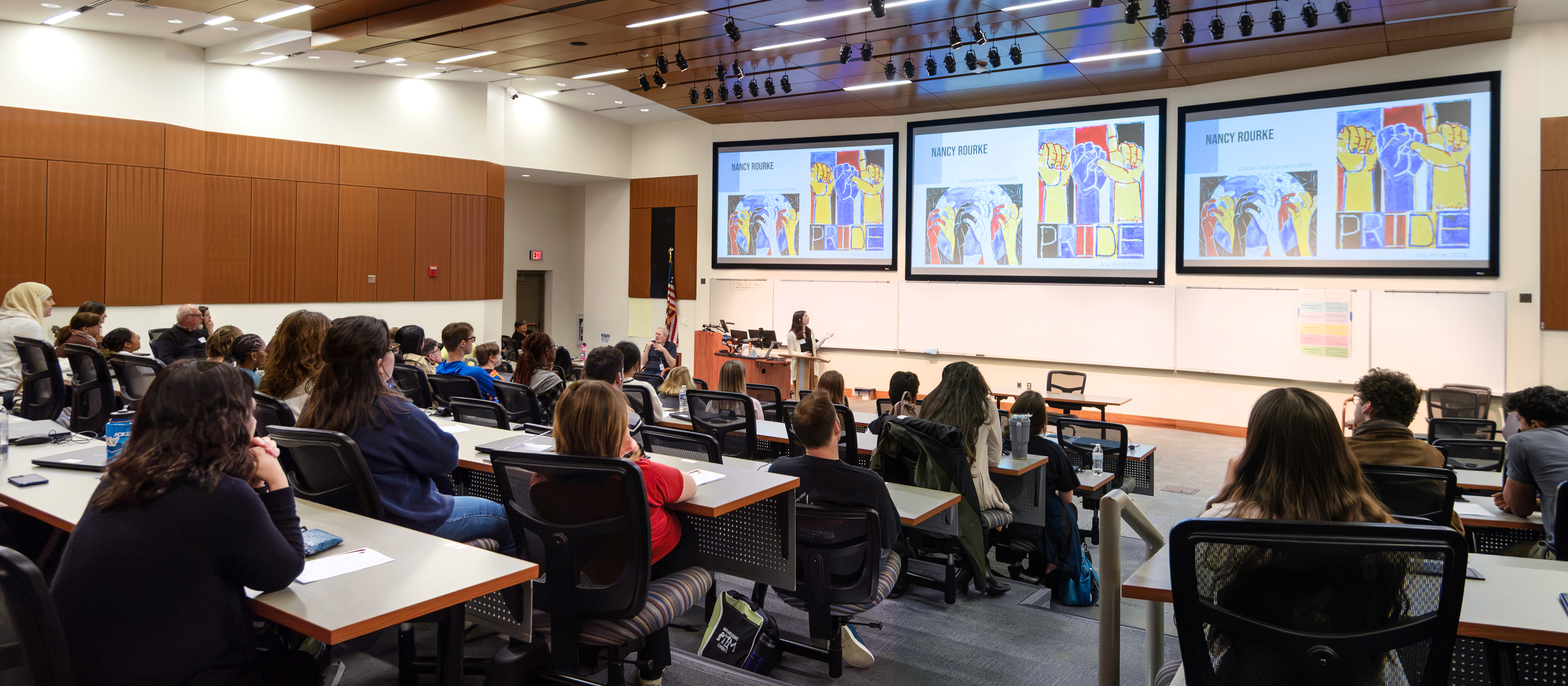 Students and faculty attend a scholarly presentation in a lecture hall, with research artwork displayed on large screens at the front.