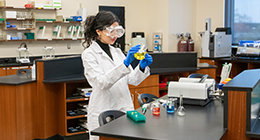 A masked student working in a lab technician wearing a white lab coat and gloves examines a blue laboratory instrument at a workbench in a science lab.