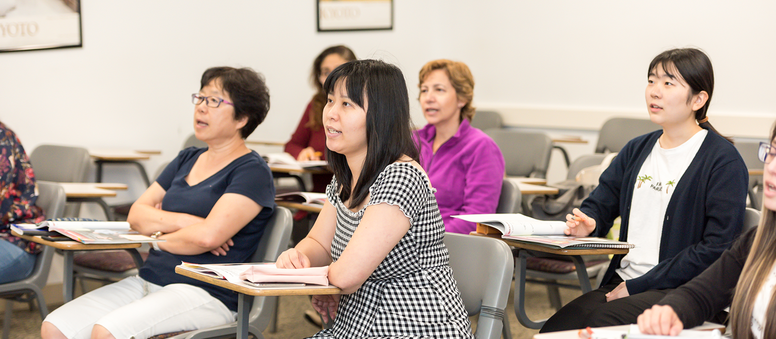 Adult students seated in a classroom, attentively listening during a lesson. Several learners sit at individual desks with open books and notebooks, facing the front of the room in an engaged, focused manner.