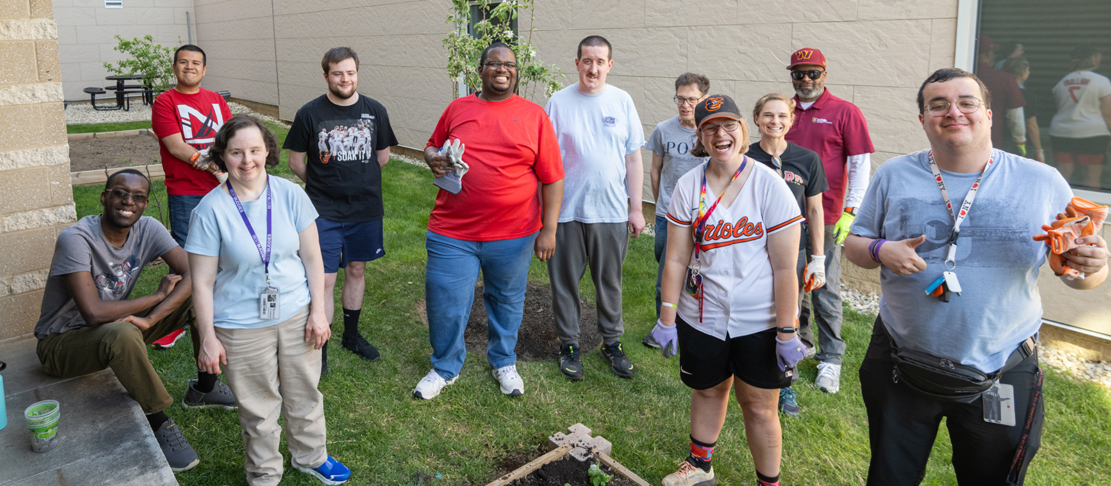 Group students smiling outdoors while gardening and planting in a courtyard beside a building. Participants wear casual clothes and gardening gloves while standing around a small raised garden bed during an outdoor activity.
