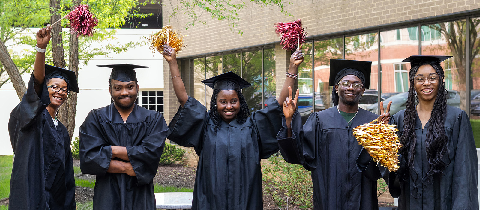 Five high school diploma graduates wearing black caps and gowns stand outdoors on HCC's campus, smiling and celebrating with maroon and gold pom-poms raised as they pose for a group photo before commencement.