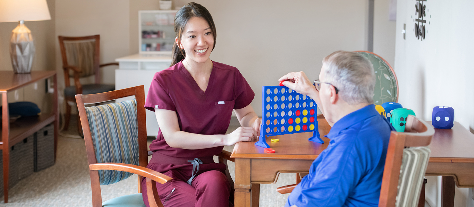 Nurse in maroon scrubs smiles while playing a tabletop game with an older adult seated across from her in a care setting.