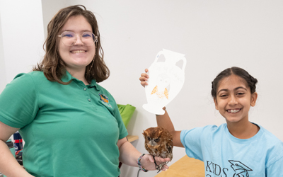 An adjunct instructor in a green shirt smiles while holding a small brown owl on her gloved hand. Next to her, a young girl wearing a light blue 