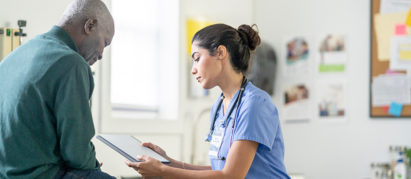 A healthcare professional in scrubs reviews information on a tablet with a seated patient in a clinical exam room.