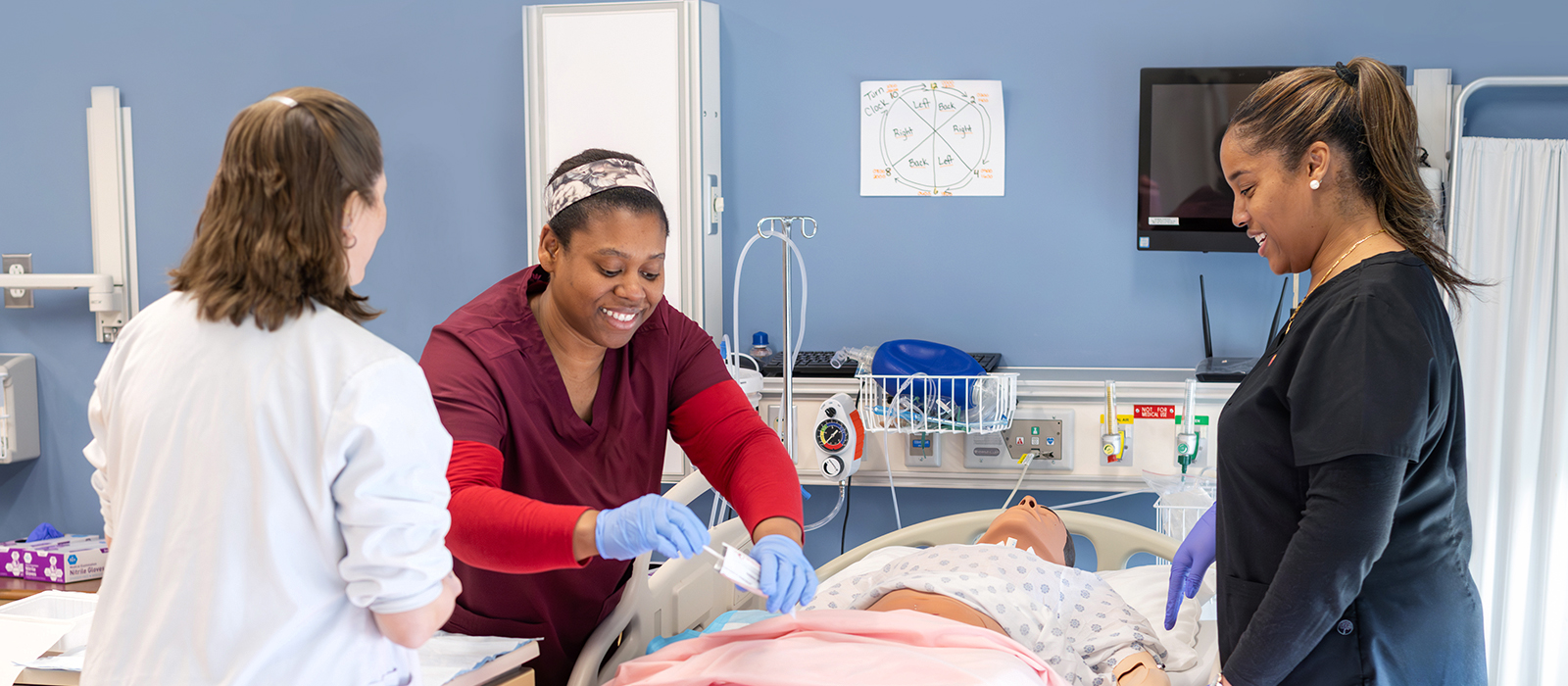 Healthcare students practice patient care in a simulated hospital room with a patient mannequin and medical equipment.