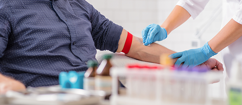 Phlebotomy student wearing gloves performs a blood draw with a tourniquet applied, with lab tubes and equipment visible in a clinical training setting.