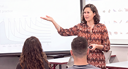 Instructor gestures toward a projected lesson while leading a classroom discussion with adult students seated at desks.